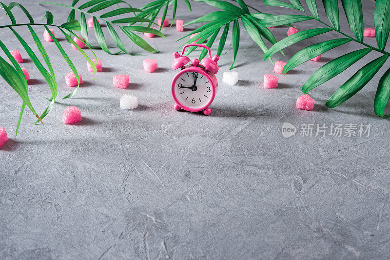 Pink alarm clock, green branches of a tropical plant and heart-shaped shapes are on a rough gray background. 情人节作文，母亲节作文。准备礼物的时间到了。